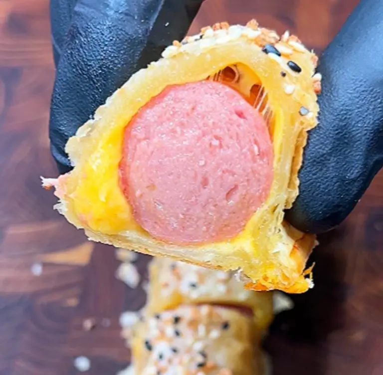 A gloved hand holds a cross-section of a pastry-wrapped sausage. The pastry is golden with a sprinkling of sesame seeds, and the sausage is pink. The background shows a wooden surface.