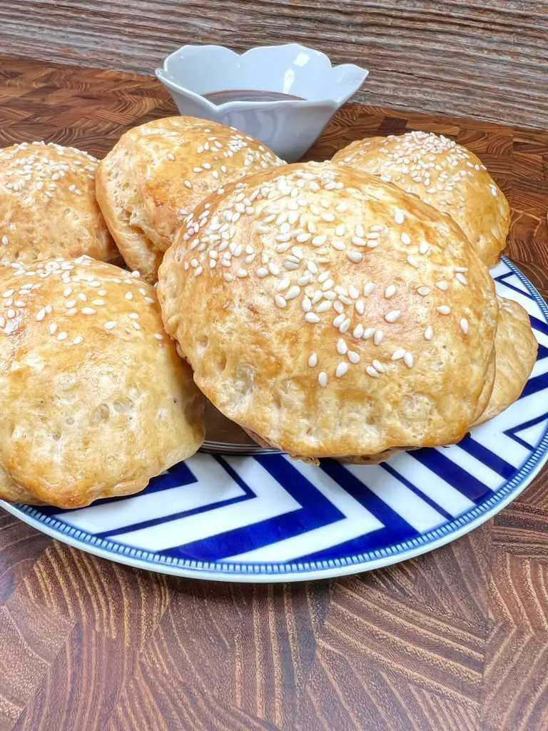 A plate with freshly baked sesame seed buns on a decorative blue and white pattern. A small, shallow bowl is in the background on a wooden surface.