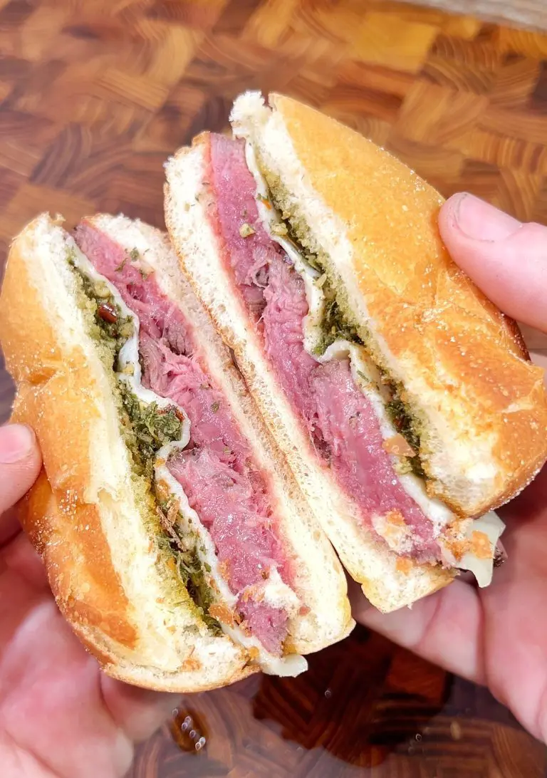 A close-up of a sandwich being held in both hands, split in half to reveal pink roast beef, cheese, and a layer of green chimichurri sauce between two halves of a toasted bun. The background is a wooden surface.