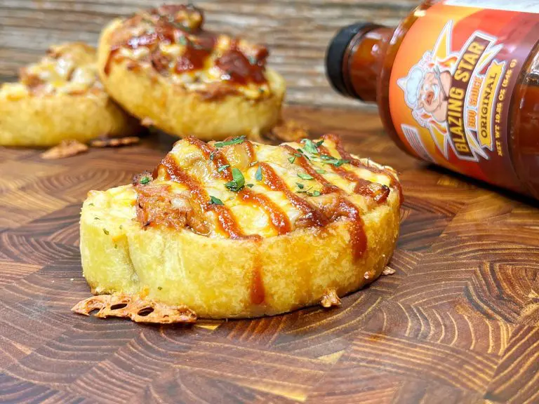 Close-up of cheesy barbecue bread topped with herbs on a wooden surface. A bottle of Blazing Star Original sauce is next to it. Two more pieces of bread are in the background.