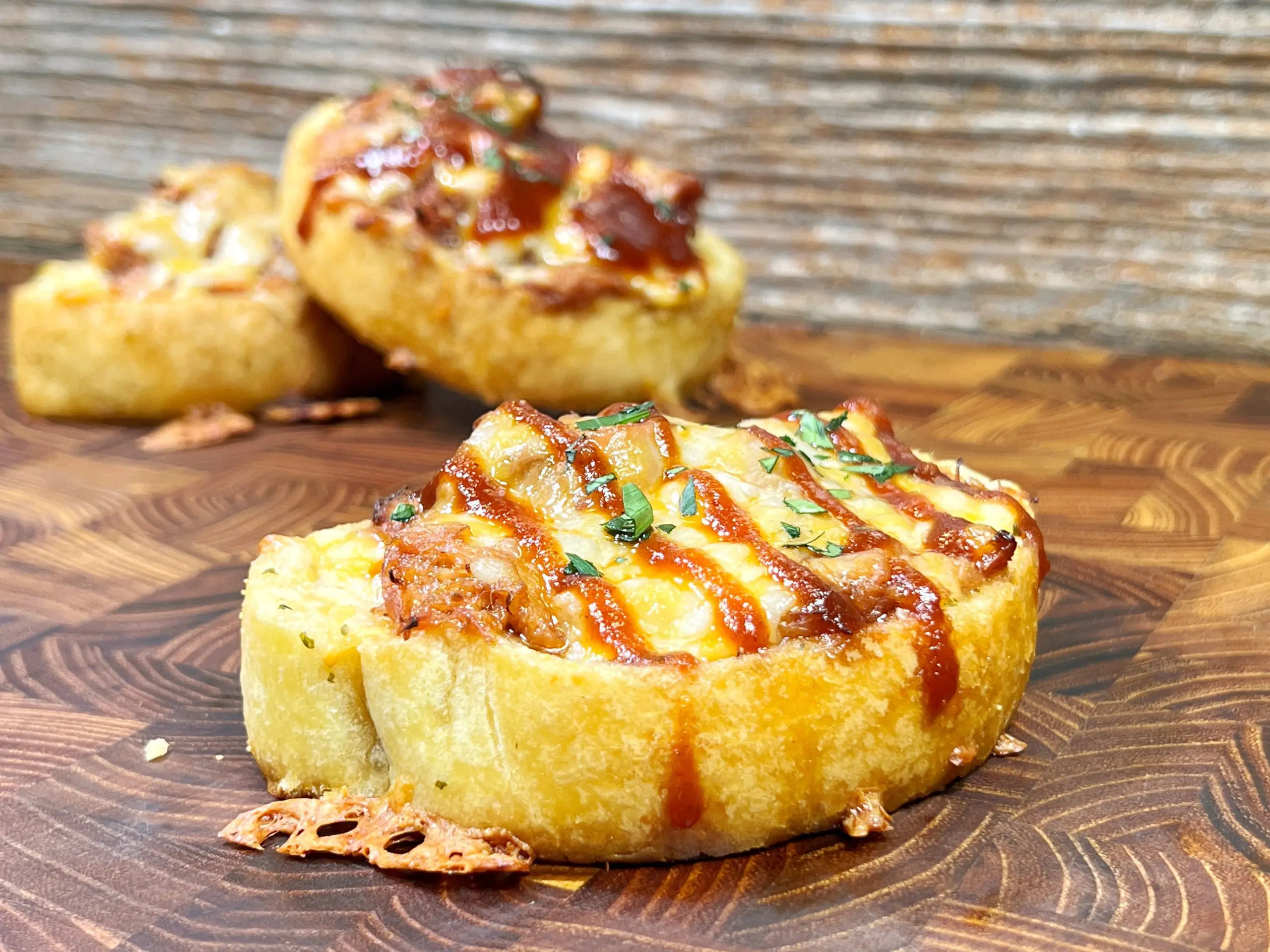 Slices of toasted bread topped with melted cheese, chopped herbs, and a drizzle of barbecue sauce are displayed on a wooden surface. The bread appears crispy and is garnished with cooked meat pieces.