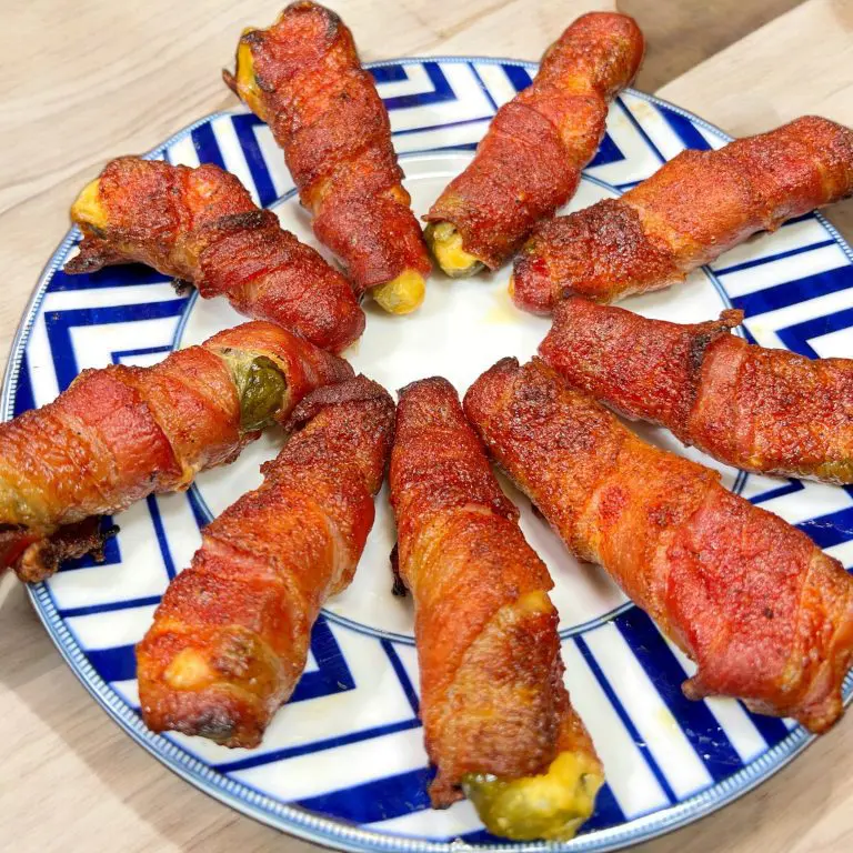 A plate with nine bacon-wrapped jalapeños arranged in a circular pattern. The jalapeños are roasted, with crispy bacon on the outside. The plate has a blue and white geometric pattern.