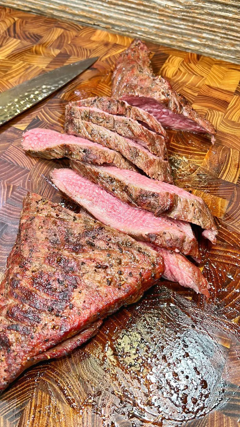 Slices of perfectly cooked medium-rare steak laid on a wooden cutting board, with a shiny, juicy texture. A carving knife is partially visible to the side. The boards rich pattern contrasts with the tender pink and brown steak.