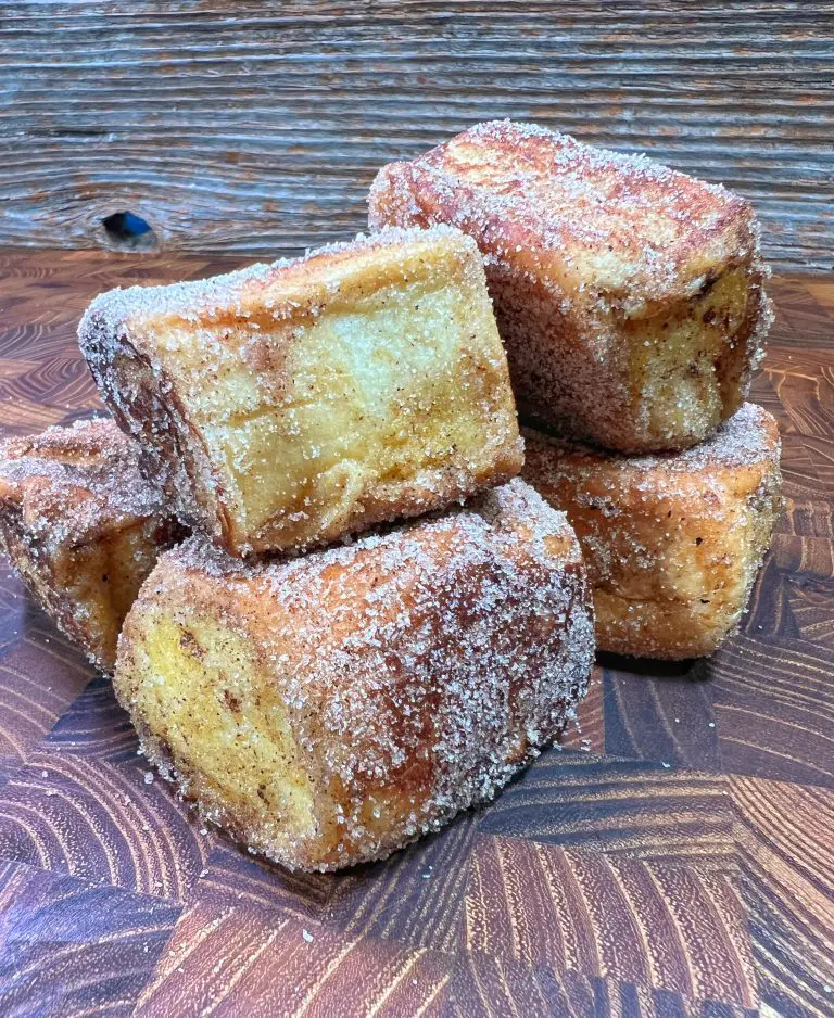 A stack of sugared pastries with a golden-brown crust sits on a wooden surface. The pastries have a rectangular shape and appear crispy, with visible layers and a coating of granulated sugar. The background features textured wood.