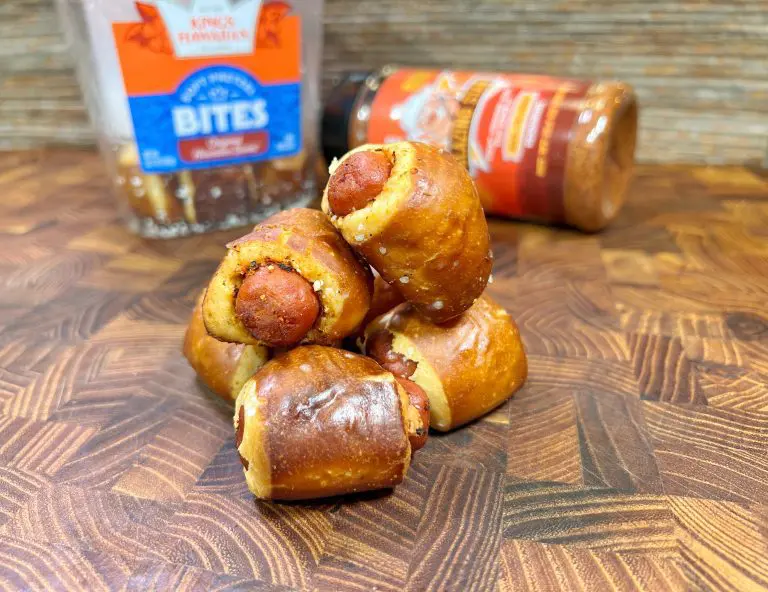 A pile of pretzel-wrapped sausages sits on a wooden surface. Behind them, there are containers of seasoning and pretzel bites. The focus is on the front pretzel-wrapped sausage, with a rustic kitchen background.