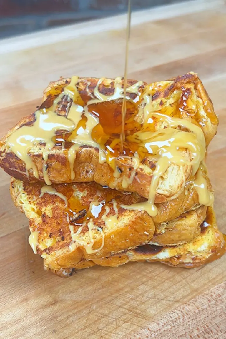 A stack of three thick slices of French toast topped with a drizzle of creamy sauce and syrup on a wooden surface. The toast is golden brown and slightly crisp on the edges.