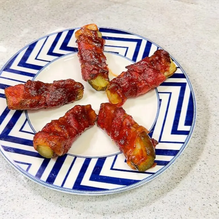 Five bacon-wrapped pickles are arranged on a decorative blue and white plate, placed on a light-colored countertop.