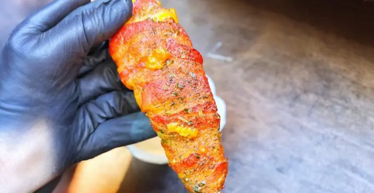 A person wearing a black glove holds a vibrant piece of food covered in red and yellow seasoning. The background shows a wooden table surface.