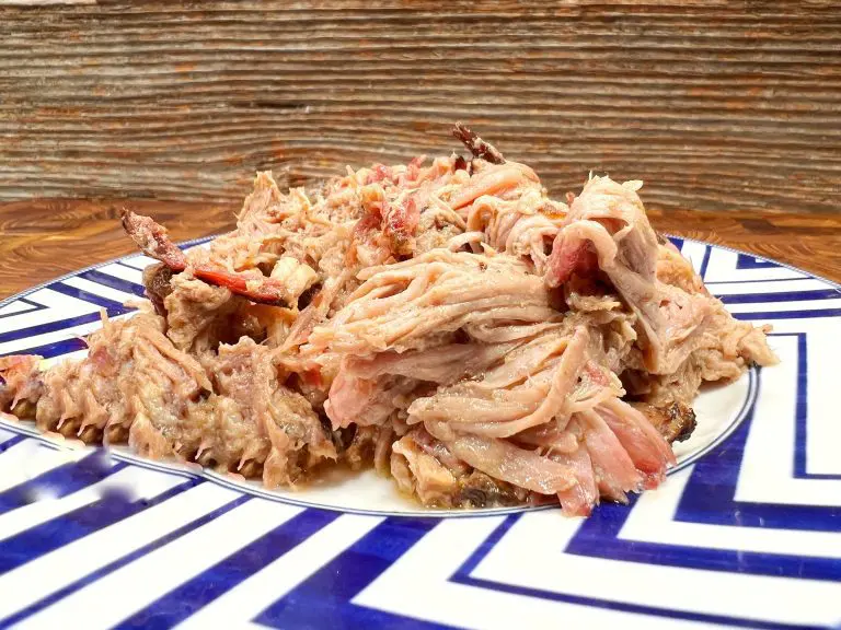 A plate of shredded pulled pork is served on a geometric blue and white patterned plate, set against a rustic wooden background. The meat appears tender and juicy.