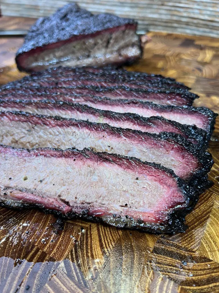 Close-up of sliced smoked brisket with a charred, blackened crust on a wooden cutting board. The tender pink interior and dark outer layers are visible in each slice, showcasing the juicy, flavorful texture that sets it apart in the pulled pork vs. brisket debate. This makes the user wonder pulled pork vs brisket