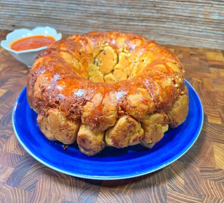 A golden-brown monkey bread on a blue plate sits on a wooden surface. A small bowl of red dipping sauce is in the background.