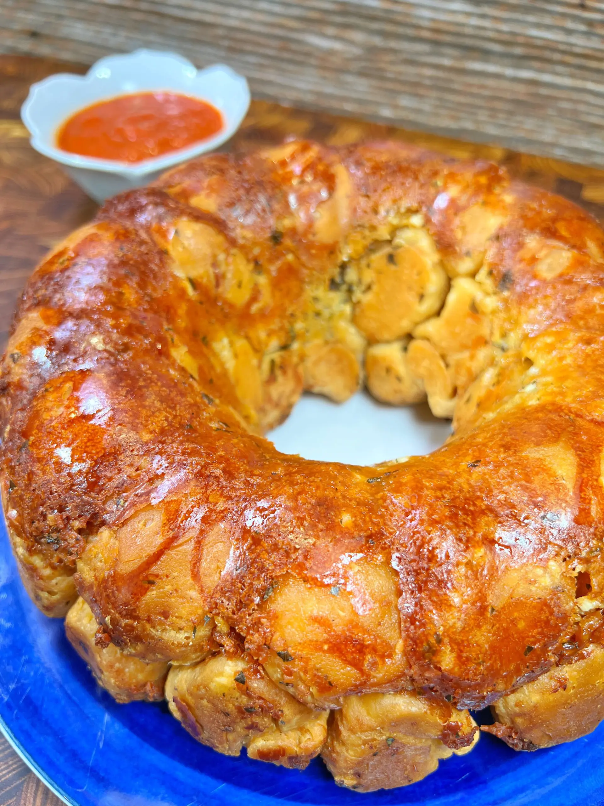 A golden-brown monkey bread sits on a blue plate, featuring a glossy, caramelized crust, with a bowl of red sauce in the background. The bread is textured and pull-apart in style, arranged in a ring shape.