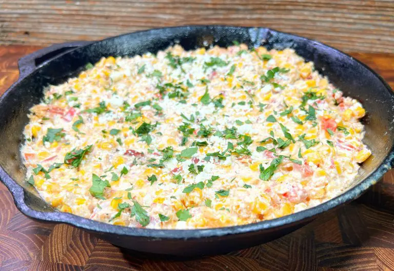 A cast iron skillet filled with creamy, cheesy corn dip topped with chopped cilantro, set on a wooden surface. The dip includes visible pieces of red and yellow peppers, creating a savory and colorful presentation.