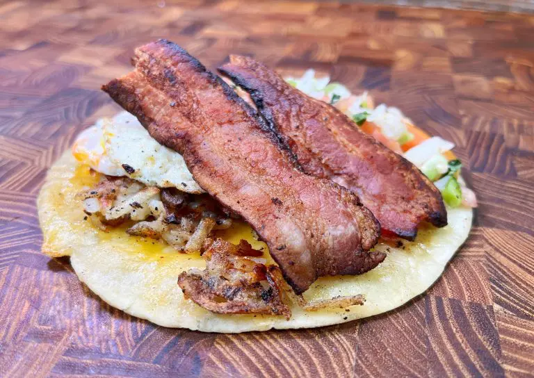 A corn tortilla topped with crispy bacon strips, a fried egg, and diced vegetables sits on a wooden surface. The bacon appears golden brown and the tortilla slightly charred.