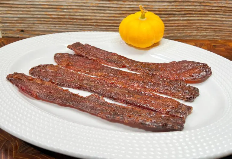 A white plate with four strips of crispy candied bacon sits on a wooden table. A small yellow pumpkin is in the background, placed against a textured wooden wall.