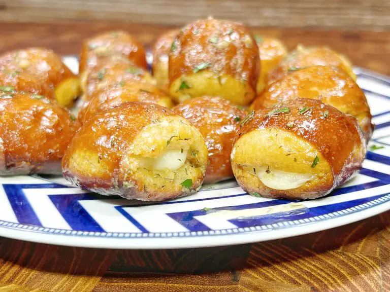 A plate of golden-brown, round pastries with a shiny glaze, garnished with herbs. Some pastries reveal a creamy filling. The plate has a blue and white geometric pattern.