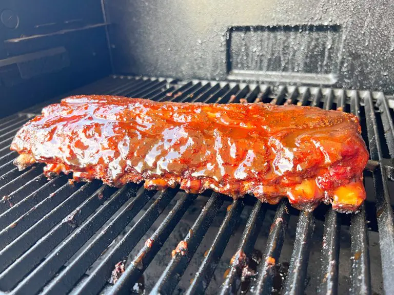 Close-up of a rack of ribs coated in barbecue sauce, cooking on a grill. The ribs are glossy and caramelized, positioned horizontally on the grill grates, with a textured metal backdrop.