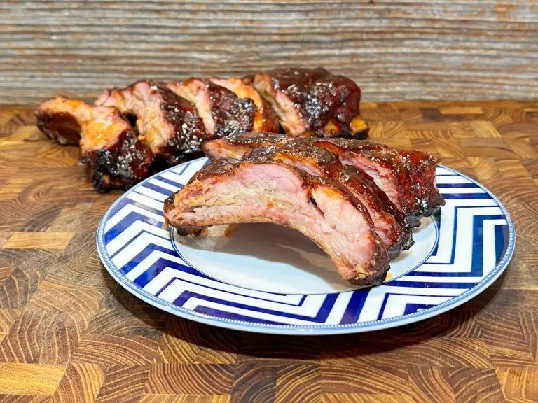 A plate of barbecued ribs with a glossy, caramelized finish on a wooden table. In the background, a larger portion of ribs is visible. The ribs are served on a plate with a blue and white zigzag pattern.