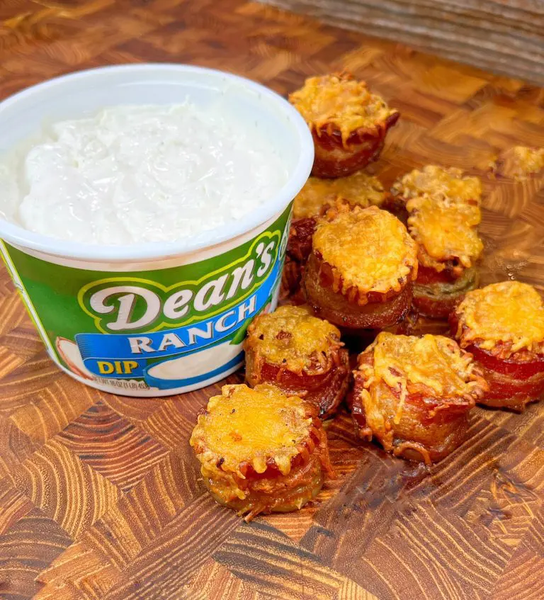 A container of Deans Ranch Dip is placed next to a pile of crispy bacon-wrapped snacks topped with melted cheese on a wooden surface.