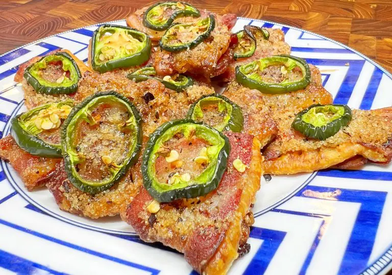 A plate of bacon-wrapped jalapeño poppers on a geometric blue and white patterned dish. The jalapeños are sliced and topped with a crispy coating, revealing the bacon underneath. The poppers are arranged on a wooden table surface.