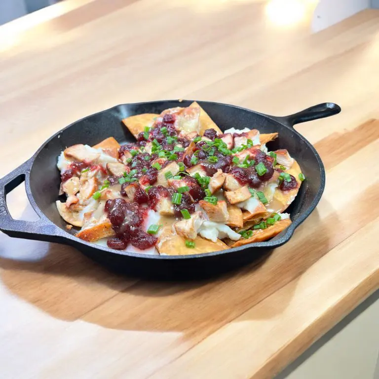 A skillet filled with nachos topped with melted cheese, diced chicken, cranberry sauce, and chopped green onions, placed on a wooden table.