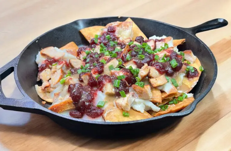 A cast-iron skillet brimming with nachos topped with Thanksgiving leftovers like turkey pieces, cranberry sauce, melted cheese, and chopped green onions rests on a wooden table.