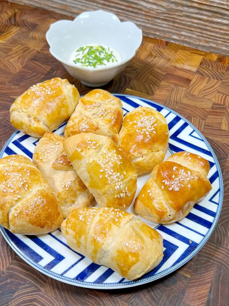 A plate of freshly baked buffalo chicken bombs topped with sesame seeds is arranged on a blue and white geometric-patterned dish. In the background, theres a small white bowl filled with a creamy dip garnished with chopped herbs. The setting is a wooden surface.