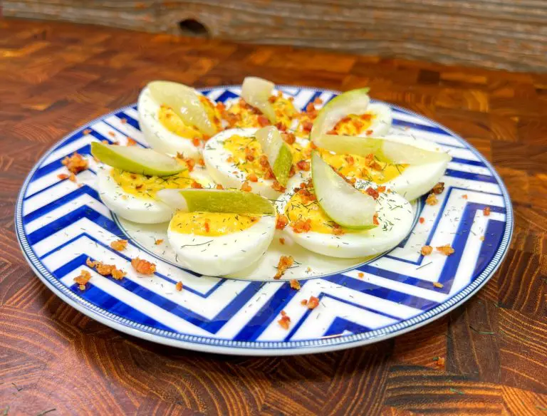 A plate of deviled eggs garnished with pear slices, dill, and crumbled bacon, arranged on a blue and white zigzag-patterned dish, placed on a wooden surface.