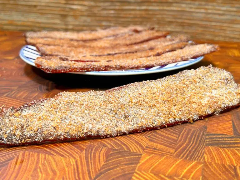 Close-up of brown sugar-coated bacon strips on a wooden surface, with more strips on a plate in the background. The bacon appears crispy and caramelized, adding a sweet twist to the savory treat.