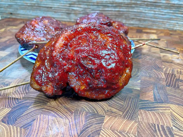 Close-up of skewered, grilled meat coated in a glossy red sauce, laid on a decorative plate with a wooden textured background.