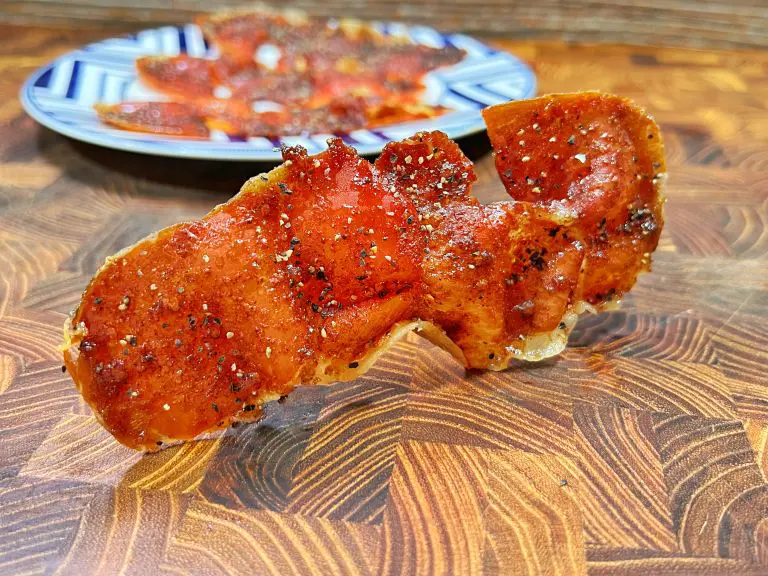 Close-up of a crispy, seasoned slice of pepperoni on a wooden surface. A plate with additional pepperoni slices is blurred in the background, emphasizing the texture and spices on the main slice.