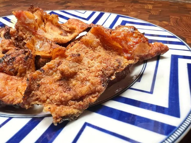 Close-up of crispy turkey skin on a plate with a blue geometric pattern. The golden-brown textures of these savory snacks stand out against the wooden table background, inviting a delicious crunch.