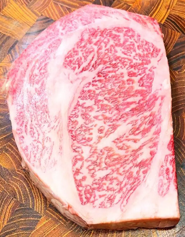 A close-up of a raw steak with rich marbling patterns, showcasing a blend of red meat and white fat. The wooden cutting board beneath contrasts with the intricate marbling.