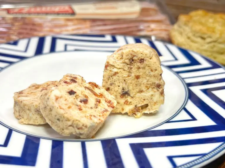 Three slices of compound butter with herbs and spices on a decorative blue and white plate. A pack of butter and a scone are blurred in the background.