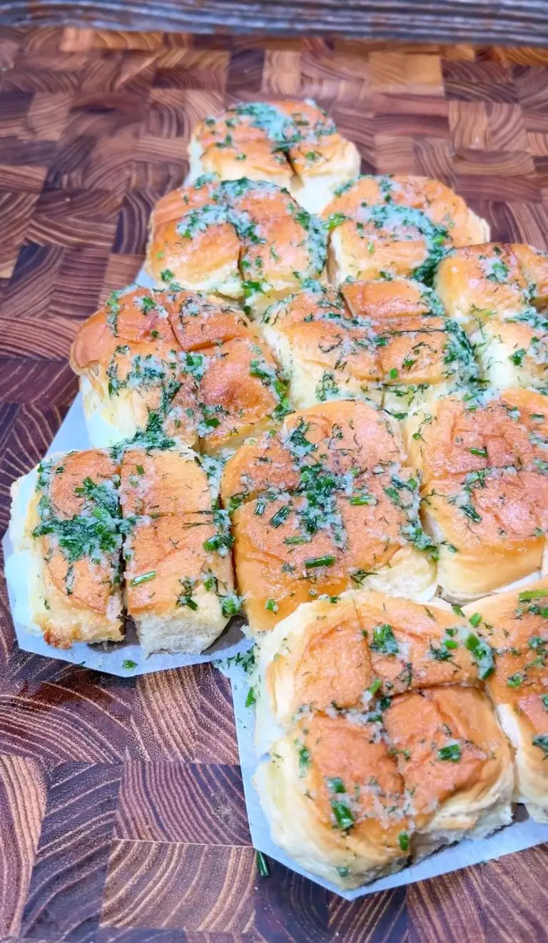 A batch of freshly baked garlic butter rolls on a wooden surface. The rolls are golden brown and topped with finely chopped herbs, glistening with garlic butter.