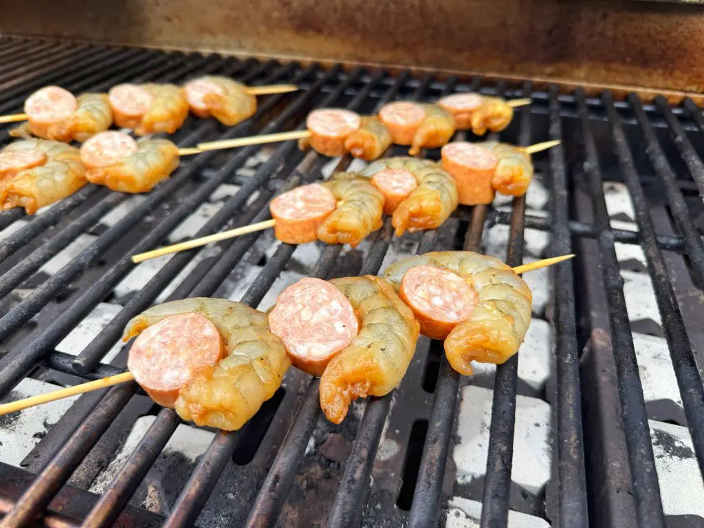 Shrimp and sausage pieces are skewered on wooden sticks and grilling on a barbecue. The skewers are diagonal, and the grill grates are visible underneath them.