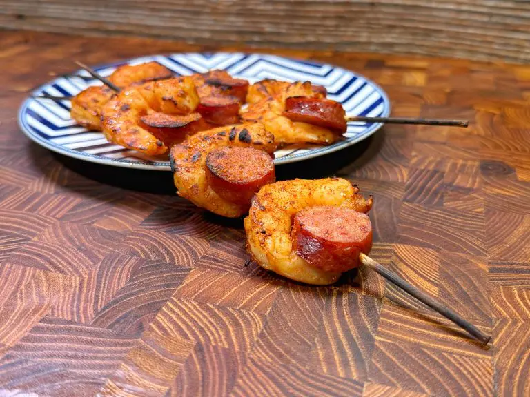 Grilled skewers of shrimp and sausage, seasoned and arranged on a white and blue patterned plate. The wooden skewers rest on a wooden surface, and the shrimp are golden brown with a charred appearance.