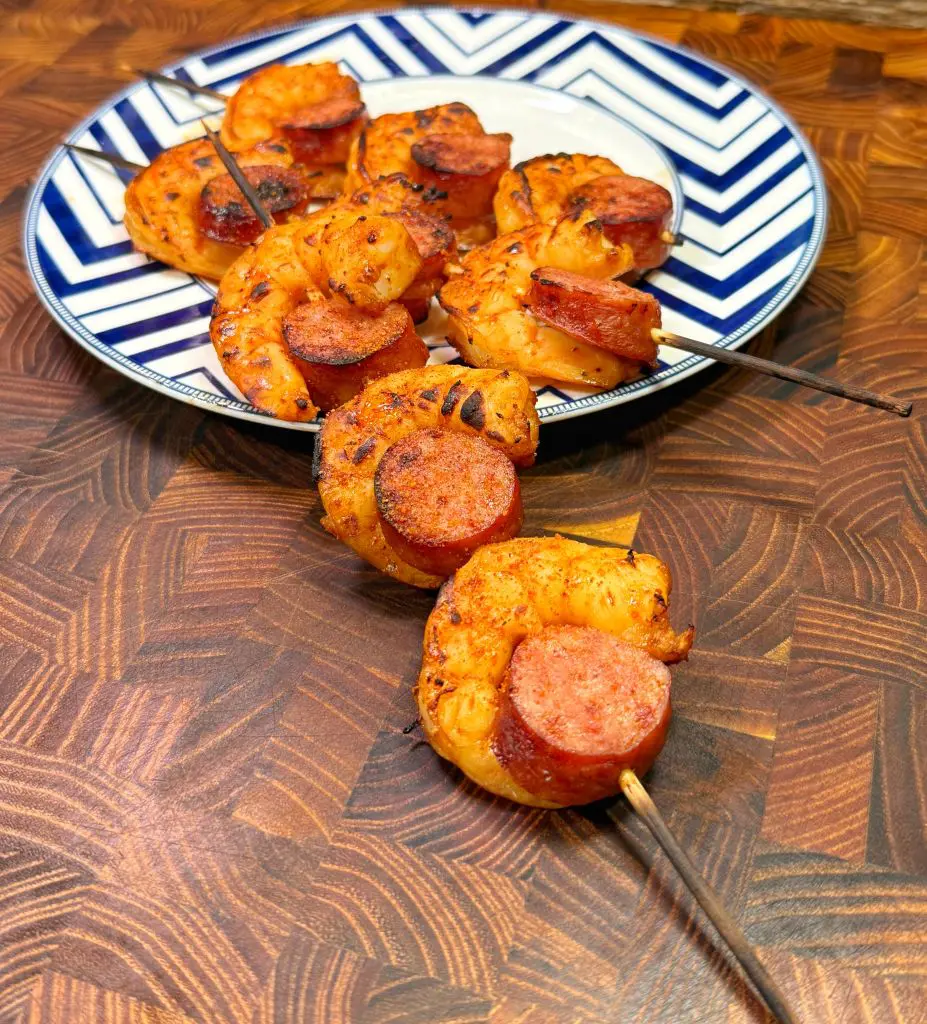 Grilled shrimp and sausage skewers on a decorative blue and white plate, placed on a wooden surface. The skewers are seasoned and cooked, showing a golden-brown texture.