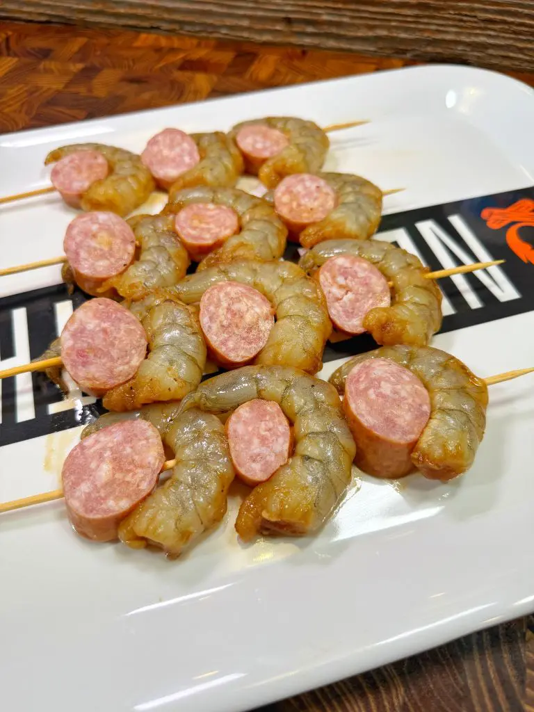 Raw shrimp and sausage slices are skewered alternately on sticks, arranged on a rectangular white plate with a decorative pattern.
