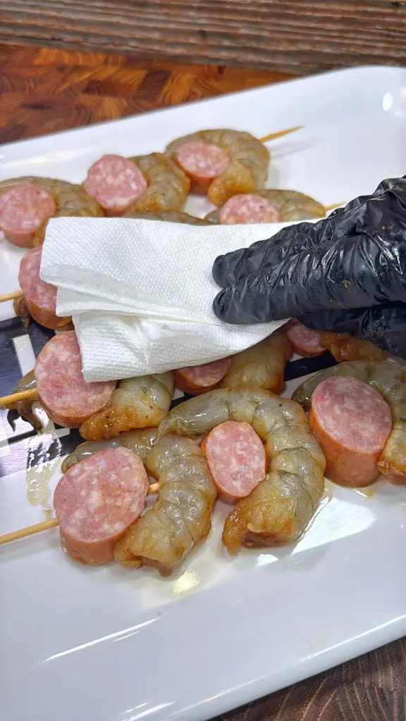 A gloved hand holding a paper towel is patting dry skewers with raw shrimp and sausage slices on a white plate. The background features a wooden surface.