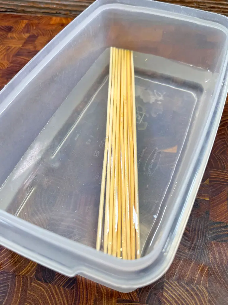 A plastic container filled with water, holding several bamboo skewers that are soaking. The container is placed on a wooden surface.