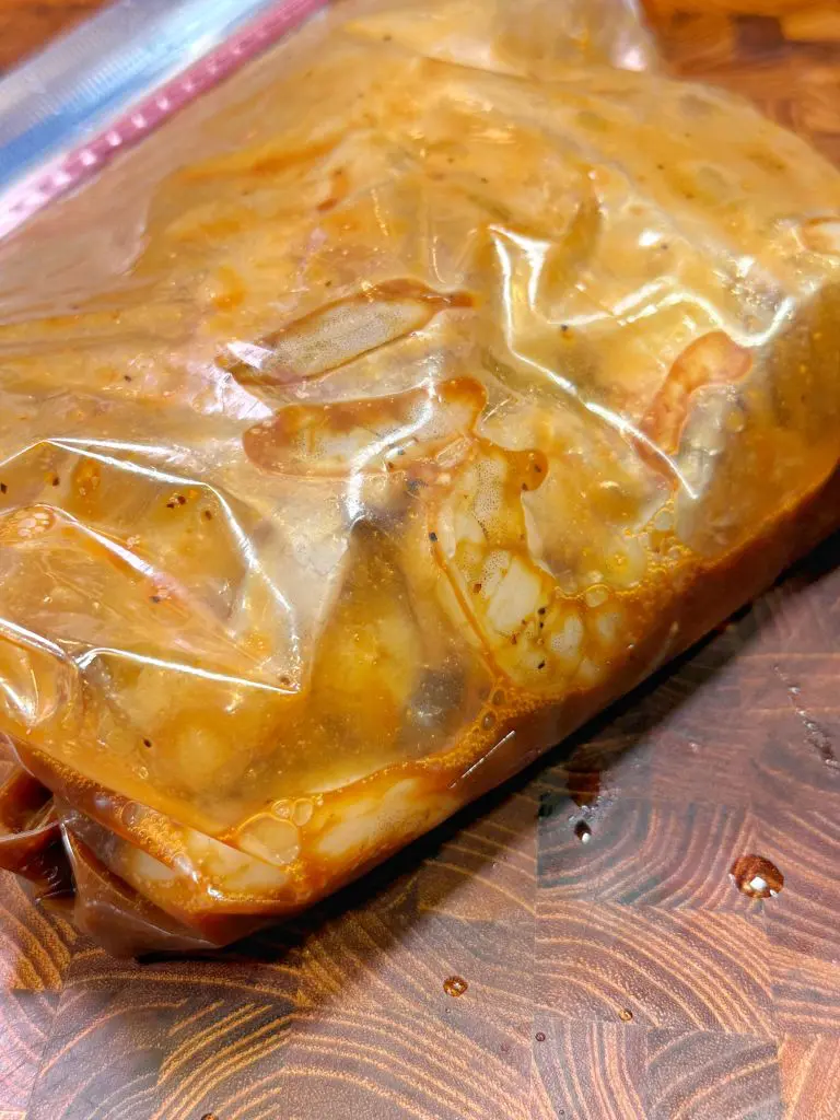 A resealable plastic bag filled with marinated chicken pieces, showing the seasonings and juices. The bag is placed on a wooden surface, with some marinade visible outside the bag.