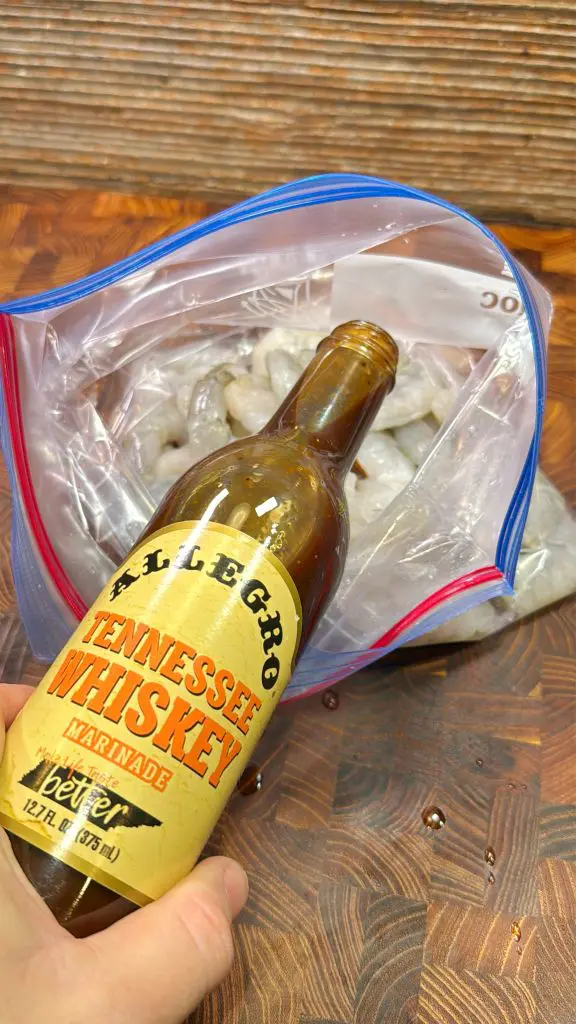 A hand pouring Allegro Tennessee Whiskey marinade into a resealable plastic bag filled with uncooked shrimp. The background features a wooden surface.