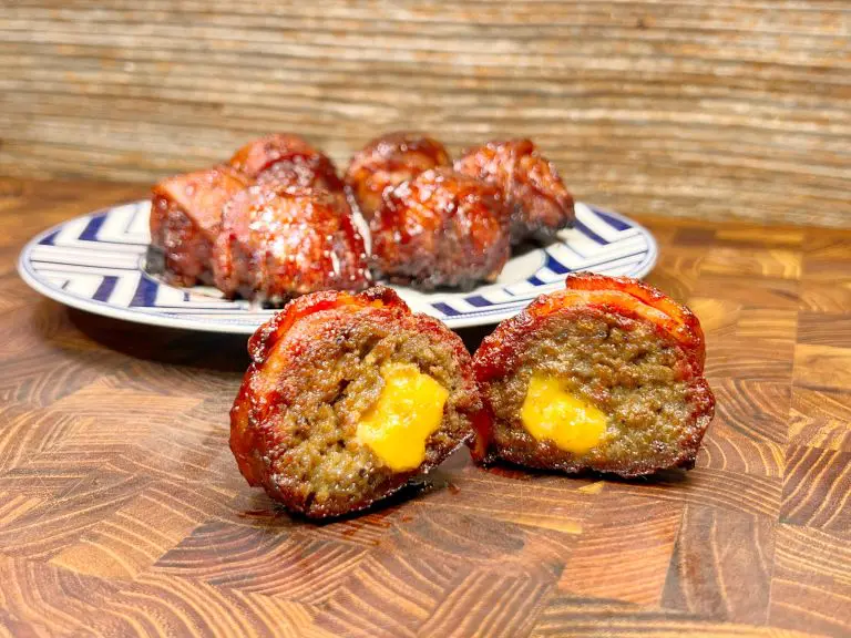 Close-up of smoked bacon-wrapped meatballs filled with melted cheese on a wooden surface. A plate with more meatballs is in the background. The texture of the meatballs and the glossy glaze are visible.