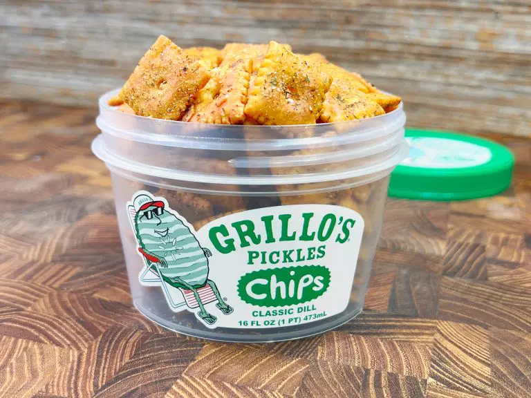 A plastic container of Grillos Pickles Chips, Classic Dill flavor, is filled with seasoned crackers. The container has a playful cartoon pickle character on the label, and a green lid rests nearby on a wooden surface.