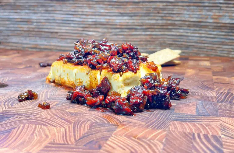 A block of cream cheese topped with a colorful mixture of chopped sun-dried tomatoes and herbs sits on a wooden cutting board. In the background, a few crackers are partially visible. The texture of the board and toppings is detailed.