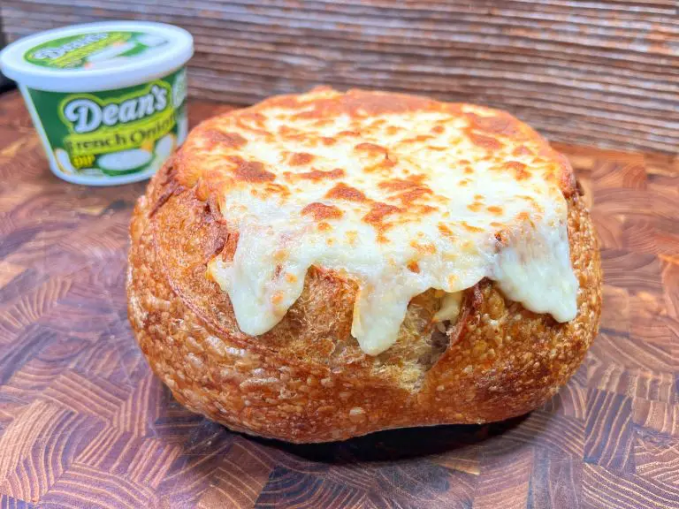 A bread bowl filled with melted cheese placed on a wooden surface. In the background, there is a container of Deans French Onion Dip. The cheese is golden and slightly bubbly, overflowing the crusty bread bowl.