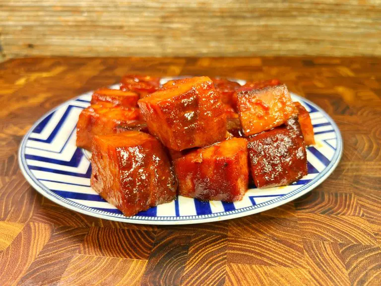 A ceramic plate with a blue and white zigzag pattern holds cubes of glazed pork belly, placed on a wooden surface. The meat appears caramelized and shiny from the sauce.