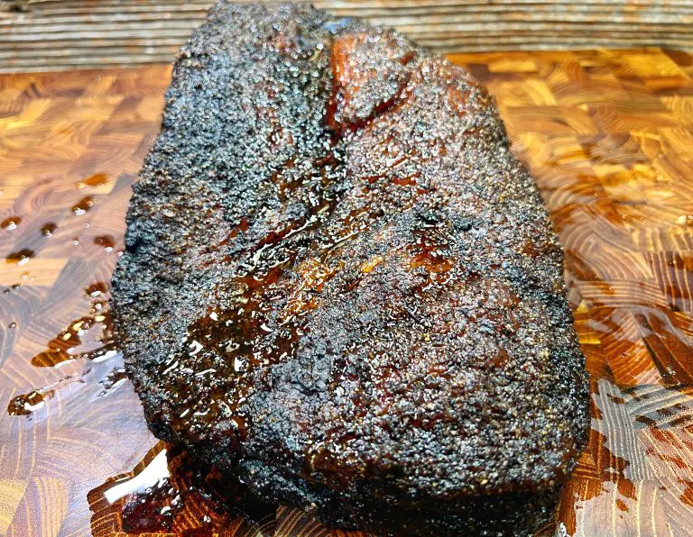 A close-up of a smoked, spice-rubbed brisket resting on a wooden cutting board. The brisket has a dark, caramelized crust and glistens with juicy moisture.