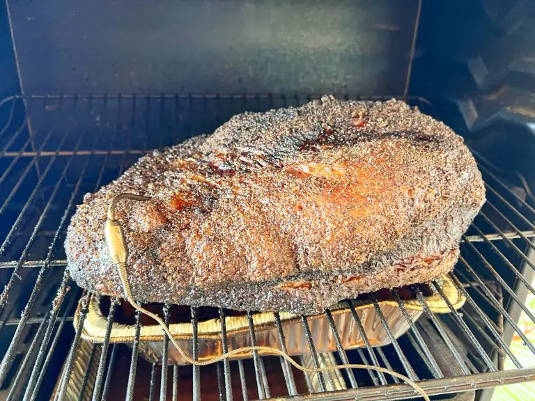 A large piece of smoked brisket with a crispy, dark crust is cooking on a grill. A meat thermometer is inserted into the brisket, and it rests on a foil tray inside the smoker.